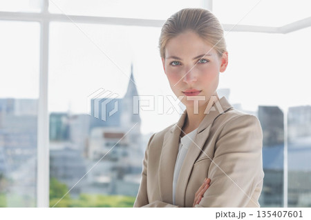 Businesswoman standing in office wearing blazer overlooking skyline by glass wall, copy space 135407601