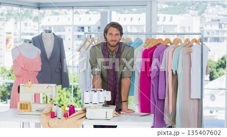 Male fashion designer leaning on white worktable in bright design studio with two sewing machines 135407602