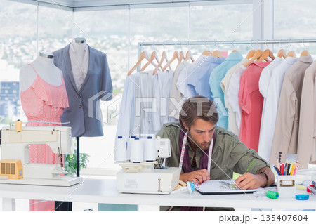 Male fashion designer sketching in notebook at design studio with sewing machines, copy space 135407604