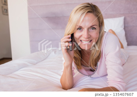 Woman lying on bed holding smartphone to ear, smiling toward camera in pink headboard bedroom Woman lying on bed holding smartphone to ear, smiling toward camera in pink headboard bedroom 135407607
