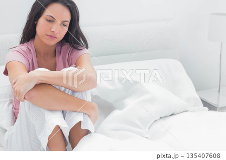 Woman sitting in pajamas on bed at home, looking downward at nightstand lamp, copy space Woman sitting in pajamas on bed at home, looking downward at nightstand lamp, copy space 135407608