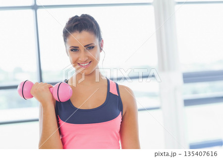 Woman smiling while wearing athletic top holding pink dumbbell at gym by window grid with earbuds 135407616