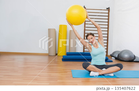 Woman lifting yellow exercise ball overhead sitting cross-legged on blue yoga mat in fitness studio Woman lifting yellow exercise ball overhead sitting cross-legged on blue yoga mat in fitness studio 135407622