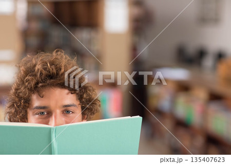 Man peeking over light green hardcover book at library with wooden shelves and tables and chairs Man peeking over light green hardcover book at library with wooden shelves and tables and chairs 135407623