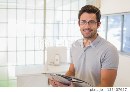 Office worker standing and smiling in office holding newspaper by glass partition, copy space Office worker standing and smiling in office holding newspaper by glass partition, copy space 135407627