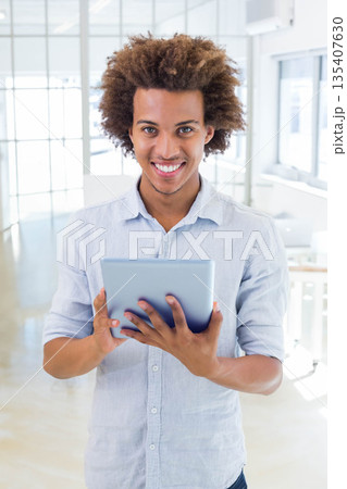 Man standing in modern office wearing light blue shirt holding tablet at chest level facing camera Man standing in modern office wearing light blue shirt holding tablet at chest level facing camera 135407630