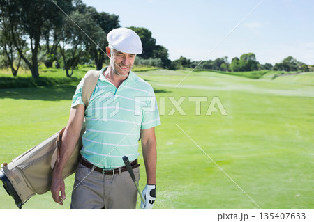 Middle-aged male golfer walking on fairway wearing striped polo shirt carrying golf bag, copy space 135407633
