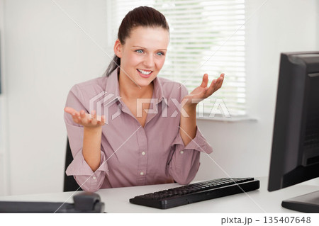 Woman wearing purple blouse smiling and gesturing toward computer monitor at office desk 135407648