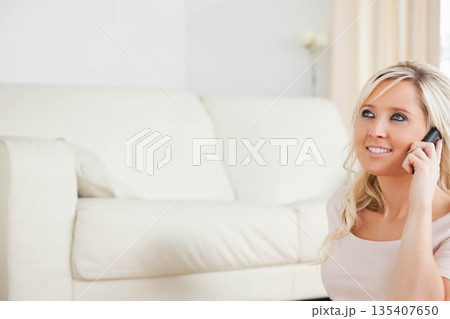 Woman with blonde hair sitting on carpet beside leather couch, talking on smartphone, copy space Woman with blonde hair sitting on carpet beside leather couch, talking on smartphone, copy space 135407650