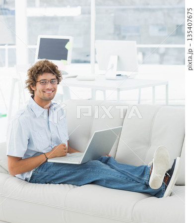 Man wearing glasses sitting on leather sofa in office using laptop near desk monitors, copy space 135407675