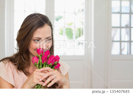 Woman in her thirties holding and smelling pink rose bouquet near face in sunlit home room Woman in her thirties holding and smelling pink rose bouquet near face in sunlit home room 135407685