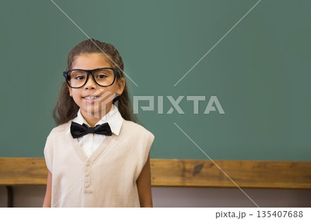 Hispanic child standing at chalkboard chalk tray in classroom wearing eyeglasses bowtie, copy space Hispanic child standing at chalkboard chalk tray in classroom wearing eyeglasses bowtie, copy space 135407688