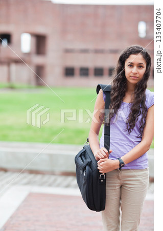 Woman standing on campus walkway holding black messenger bag and checking wristwatch, copy space 135407704