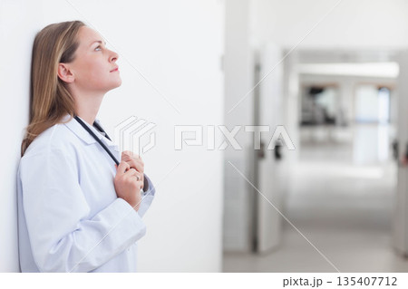 Mid adult female doctor standing by white wall in corridor wearing coat and stethoscope, copy space 135407712