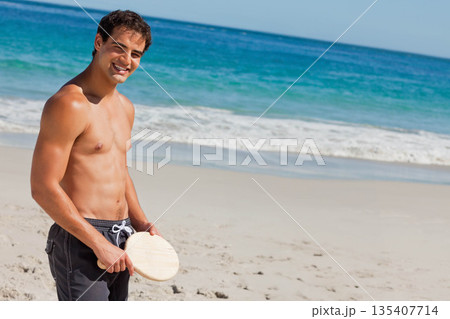 Shirtless man wearing swim trunks holding wooden paddle and standing on shore by waves, copy space Shirtless man wearing swim trunks holding wooden paddle and standing on shore by waves, copy space 135407714