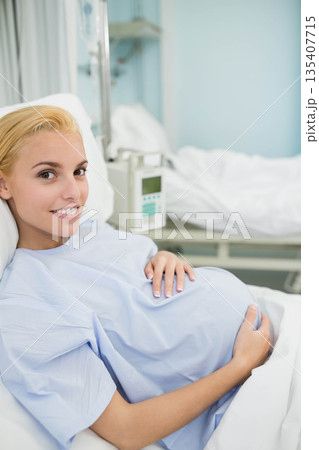 Pregnant woman holding belly reclining on hospital bed wearing gown beside IV stand Pregnant woman holding belly reclining on hospital bed wearing gown beside IV stand 135407715