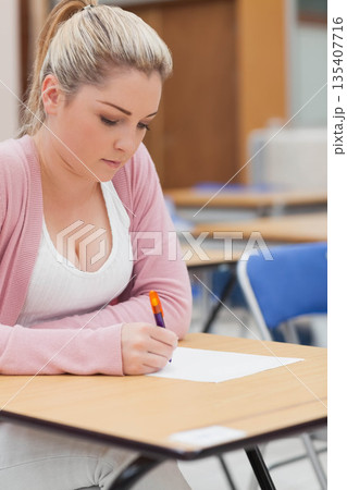 Female student sitting on blue chair at desk in classroom writing on paper with orange pen 135407716