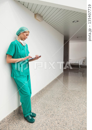 Female nurse in scrubs with mask leaning against wall using tablet in hospital corridor, copy space Female nurse in scrubs with mask leaning against wall using tablet in hospital corridor, copy space 135407719