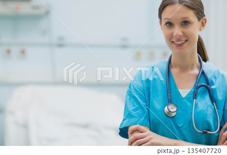 Female nurse standing with arms crossed wearing blue scrubs in hospital room near bed, copy space 135407720