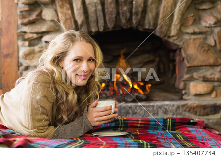 Woman reclining on plaid blanket by stone fireplace in cabin holding ceramic mug, copy space 135407734
