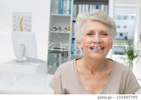 Senior female sitting and smiling in clinic office with monitor, spine chart, plant, copy space Senior female sitting and smiling in clinic office with monitor, spine chart, plant, copy space 135407745