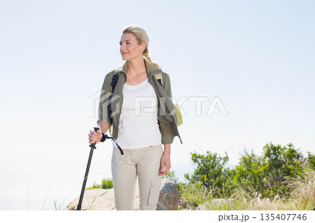 Woman gazing at horizon on rocky hillside wearing backpack with sleeping mat gripping trekking pole 135407746