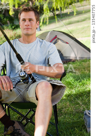 Man holding fishing rod while sitting on foldable camping chair at grassy campsite near dome tent 135407755