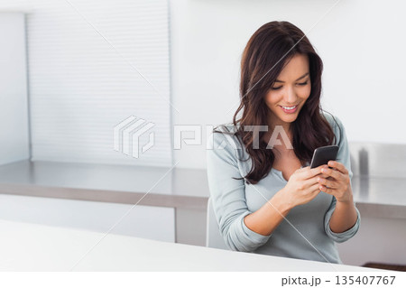 Woman smiling at smartphone while sitting at kitchen counter by shutter cabinet door, copy space Woman smiling at smartphone while sitting at kitchen counter by shutter cabinet door, copy space 135407767
