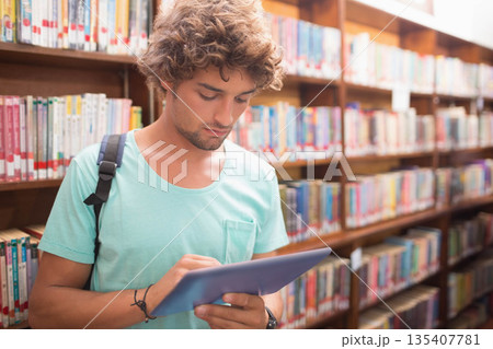 Man standing between wooden bookshelves in library aisle scanning tablet screen wearing backpack 135407781