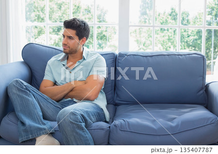 Man in mid-30s sitting cross-legged on blue sofa in living room near window with green foliage Man in mid-30s sitting cross-legged on blue sofa in living room near window with green foliage 135407787