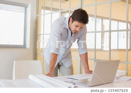 Man in button-down shirt reviewing blueprints on white desk with silver laptop at office Man in button-down shirt reviewing blueprints on white desk with silver laptop at office 135407829