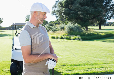 Senior man wearing flat cap gripping golf club on golf course beside golf cart, copy space 135407834