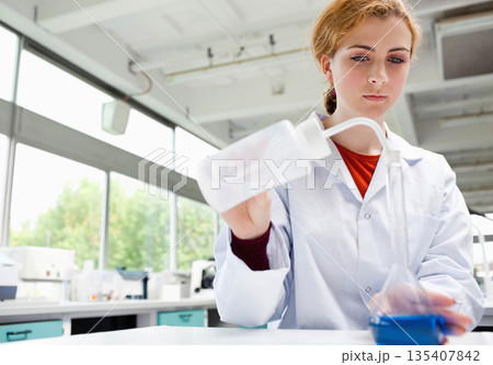 Female scientist holding white wash bottle, pouring into blue beaker at lab bench, copy space 135407842
