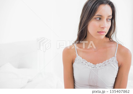 Woman sitting on edge of bed in bright white bedroom wearing lace-trimmed camisole, copy space Woman sitting on edge of bed in bright white bedroom wearing lace-trimmed camisole, copy space 135407862