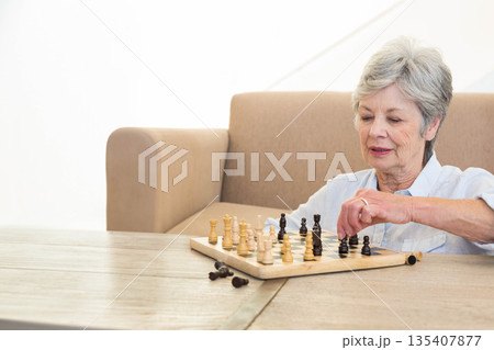 Senior woman focusing on chessboard while playing chess on wooden table in living room, copy space 135407877