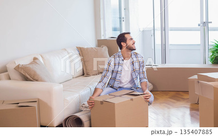 Man looking toward balcony doors while sitting on cardboard moving box in living room Man looking toward balcony doors while sitting on cardboard moving box in living room 135407885