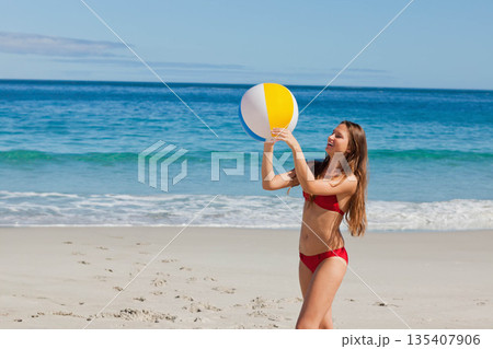 Woman standing wearing red bikini at shoreline beach holding beach ball above head, copy space Woman standing wearing red bikini at shoreline beach holding beach ball above head, copy space 135407906