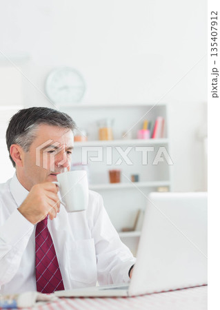 Middle-aged man holding white coffee mug, checking laptop at desk in home office, copy space 135407912