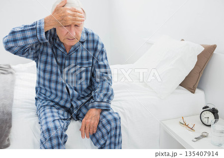 Senior man wearing plaid pajamas sitting on edge of bed holding forehead in bedroom, copy space Senior man wearing plaid pajamas sitting on edge of bed holding forehead in bedroom, copy space 135407914