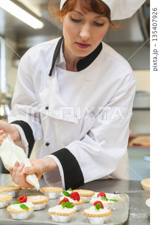 Female chef using pastry bag piping cream onto mini tarts at kitchen countertop with raspberries 135407926