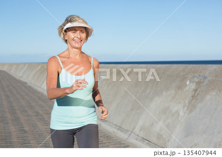 Senior woman jogging along promenade by sea wall wearing light blue tank top with sun visor Senior woman jogging along promenade by sea wall wearing light blue tank top with sun visor 135407944