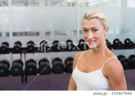 Woman standing in gym wearing white tank top and leggings by dumbbell rack, copy space Woman standing in gym wearing white tank top and leggings by dumbbell rack, copy space 135407948