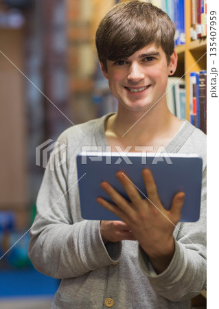 Male standing between wooden bookshelves in library aisle holding tablet with cover 135407959
