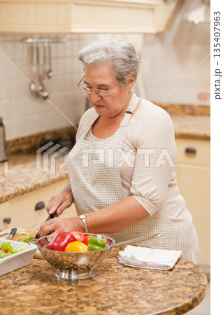 Senior woman chopping colorful peppers on cutting board on granite kitchen island with colander 135407963