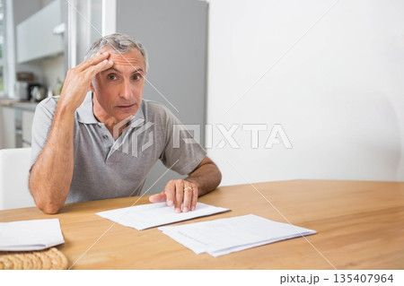 Mature male sitting at dining table reviewing paperwork with kitchen appliances visible, copy space 135407964