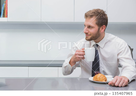 Male office worker sitting at white kitchen counter holding coffee mug and reaching for croissant Male office worker sitting at white kitchen counter holding coffee mug and reaching for croissant 135407965
