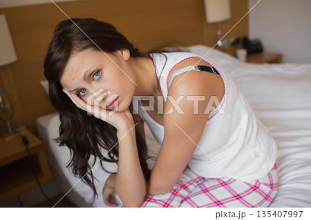 Woman wearing pajamas sitting on bed in bedroom by bedside telephone resting cheek on hand Woman wearing pajamas sitting on bed in bedroom by bedside telephone resting cheek on hand 135407997