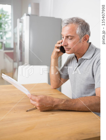 Senior man holding smartphone while reviewing printed documents with pen at kitchen dining table Senior man holding smartphone while reviewing printed documents with pen at kitchen dining table 135407998