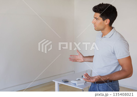 Man gesturing beside white table with telephone remote keyboard in empty room, copy space 135408021