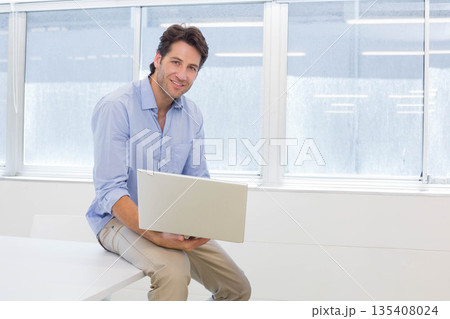 Man working on silver laptop while sitting at white desk in modern office wearing business attire 135408024